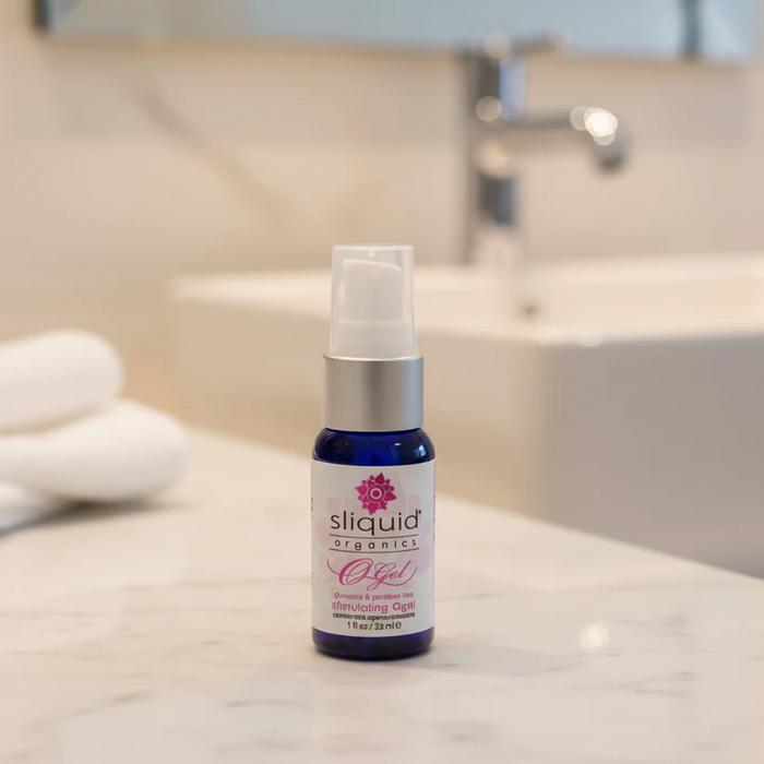 A 1 oz bottle of Sliquid Organics Stimulating Clitoral Orgasm Gel rests on a bathroom counter, with a sink, faucet, and neatly folded white towels blurred in the background.