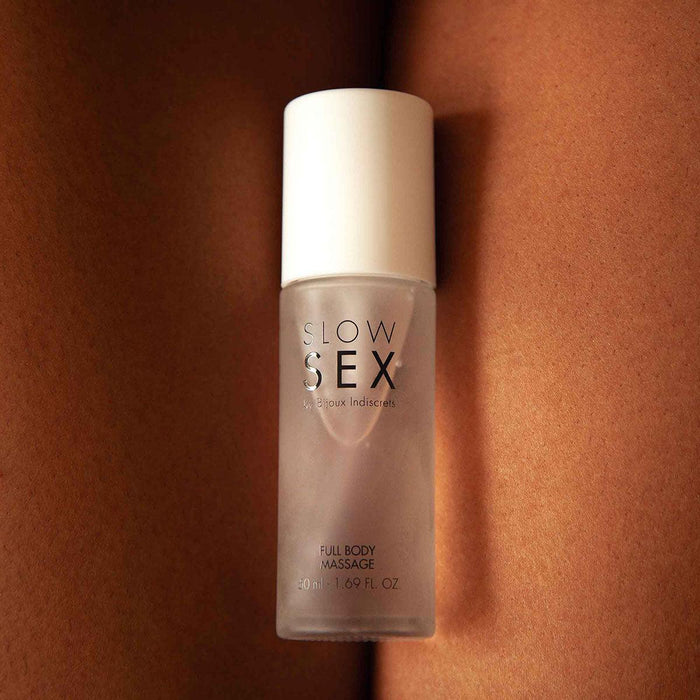 A bottle of "Slow Sex Full Body Massage Gel" by Bijoux Indiscrets, infused with a coconut scent, is placed upright on a surface with a peach-toned, flesh-like texture in the background. The sleek, semi-transparent bottle with a white cap promises body-on-body fun. The text on the bottle is clearly visible.
