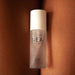 A bottle of "Slow Sex Full Body Massage Gel" by Bijoux Indiscrets, infused with a coconut scent, is placed upright on a surface with a peach-toned, flesh-like texture in the background. The sleek, semi-transparent bottle with a white cap promises body-on-body fun. The text on the bottle is clearly visible.
