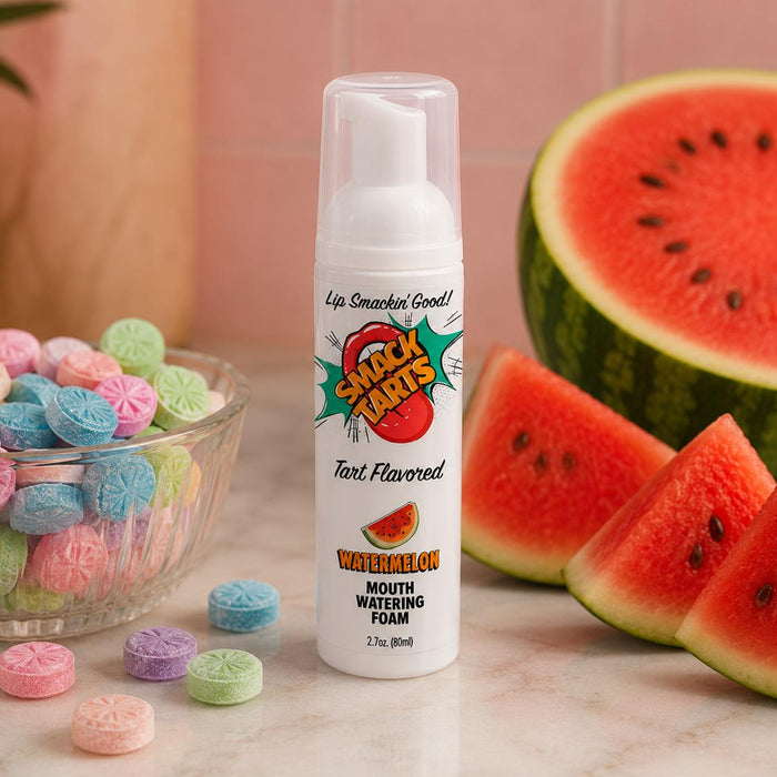 A bottle of SMACK Tarts Mouthwatering Sour Foam 2.7 oz sits on a counter among colorful candies in a glass bowl, fresh watermelon slices, and a halved watermelon in the background. Available in 6 flavors.