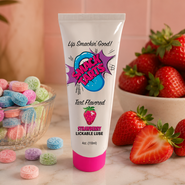 A tube of SMACK Tarts Lickable Sour Lubricant, available in 5 flavors, stands on a counter with colorful candies and strawberries, set against a pink background with a bowl of strawberries nearby.