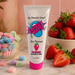 A tube of SMACK Tarts Lickable Sour Lubricant, available in 5 flavors, stands on a counter with colorful candies and strawberries, set against a pink background with a bowl of strawberries nearby.