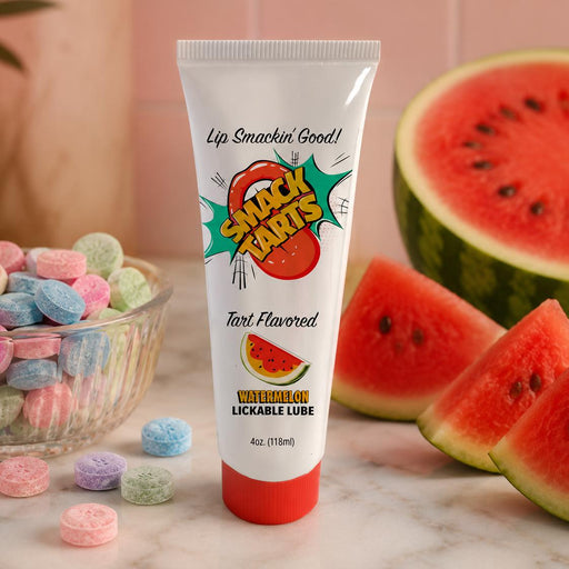 A tube labeled "SMACK Tarts Lickable Sour Lubricant" sits on a marble counter beside watermelon slices and colorful candy tablets. The tube, featuring cartoon watermelon art and comic-style text, is water-based and available in 5 flavors.