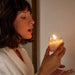 A woman in a white bathrobe gently holds the Soft Touch Massage Oil Candle by Dame (Benzoin, Guaiac Wood & Sandalwood) near her face, gazing calmly and intently at its flame.