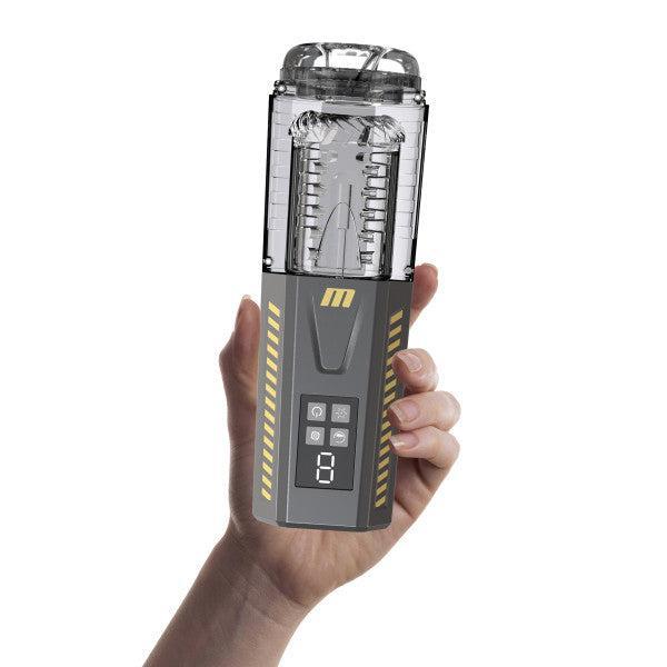 A hand holding the Blush Spin Master Pro, a futuristic and sleek gray and yellow automatic stroker with a transparent top that reveals its internal mechanisms. The front panel features a digital screen, control buttons, and an "M" logo. The device is cylindrical at the top and rectangular at the bottom, offering various vibration modes for an enhanced experience.