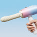 A hand holds the Spinel App-Controlled, Multi-Attachment Thrusting, Vibrating & Heating Dildo Machine with a beige, textured attachment against a light blue background.