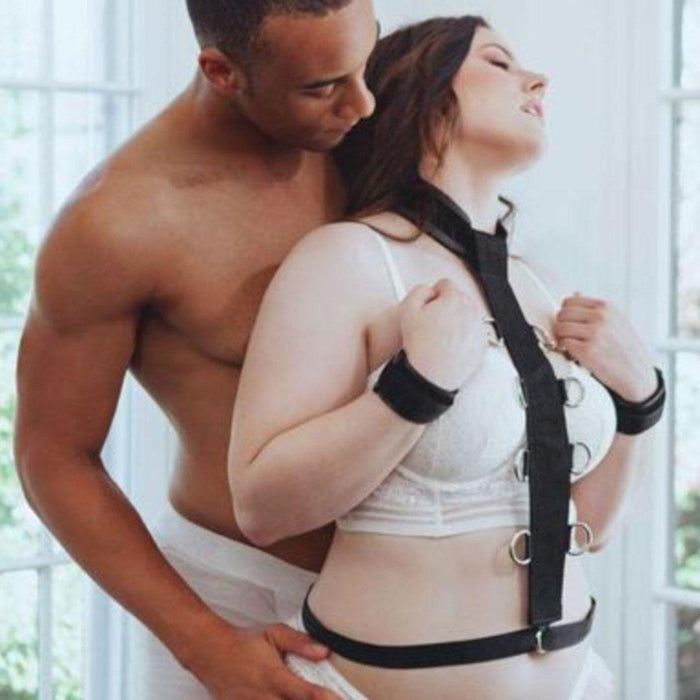 A couple sharing an intimate moment, with the man embracing the woman from behind near a bright window, both dressed in white undergarments, connected by a Sportsheets Neck and Wrist Restraint.