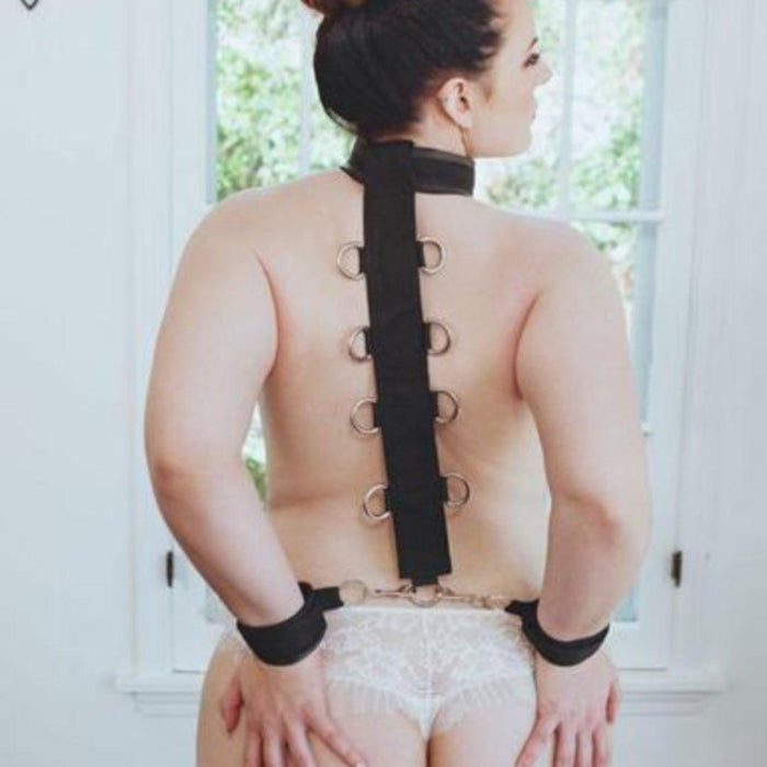 A woman wearing a Sportsheets Neck and Wrist Restraint and elegant white lingerie, sitting with her back to the camera, in a classic and serene room setting, secured by a body restraint system.