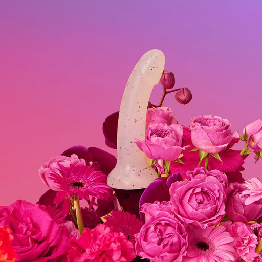 A Lovely Planet Starlight Pink Glitter 7 inch Silicone Dildo, featuring a white speckled pastel pink design and made from body-safe silicone, is positioned upright amidst a vibrant arrangement of pink and magenta flowers, including roses and gerbera daisies, against a gradient pink and purple background.
