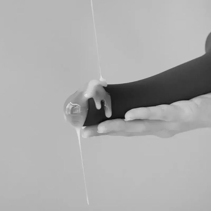 A close-up of a hand holding the Strap-On-Me 7.5 Inch Silicone Squirting Cum Dildo - Black by Lovely Planet, being drizzled with a white liquid from above. The liquid cascades over the top and sides of the dildo, reminiscent of realistic lubricant. The background is out of focus and appears to be neutral gray.
