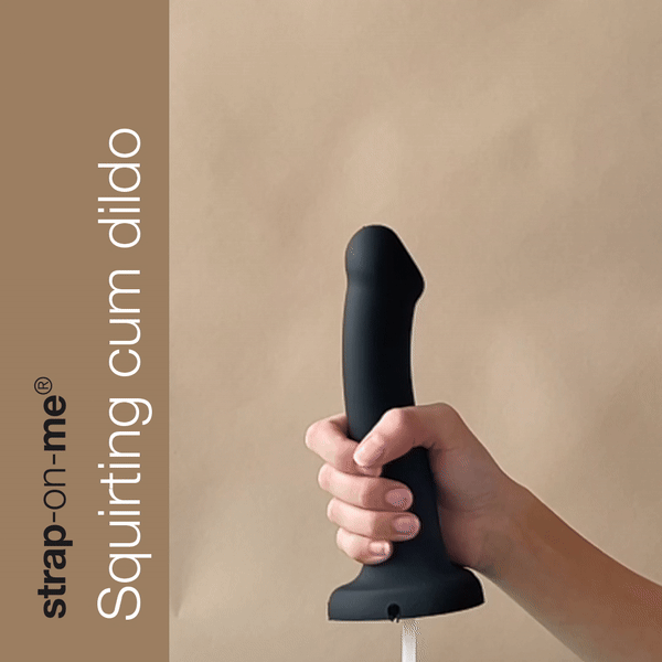 A hand holds the Strap-On-Me 7.5 Inch Silicone Squirting Cum Dildo in black, featuring a realistic design ideal for strap-on play. On the left side, text reads "Lovely Planet's strap-on-me® Squirting cum dildo." The plain beige background highlights its body-safe appeal.