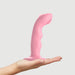 A hand is holding the Strap on Me G-Spot Tapping Silicone Dildo - Pink by Lovely Planet, showcasing its curved design and ergonomic shape with a suction base. The plain, neutral background highlights the toy's smooth texture, emphasizing its design for intimate use.