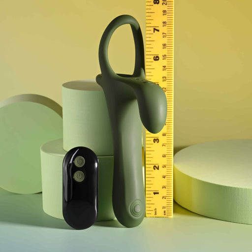 The Strap Tap & Go Girth Enhancer & Tapping Vibrator, a green silicone cock ring with a curved attachment, stands upright beside a yellow ruler showing its 6-inch height. A black two-button remote lies nearby. Circular shapes appear in the background.