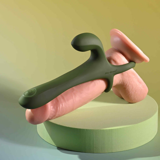 The Strap Tap & Go Girth Enhancer & Tapping Vibrator—a realistic dildo with a green strap-on harness, attached vibrating perineum stimulator, and remote control—displayed on a circular pedestal against a gradient background.