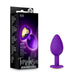 Packaging and product of the Temptasia Bling Small Silicone Butt Plug - Purple by Blush. The package on the left showcases product details, highlighting the heart-shaped jewel feature. On the right, you can see the purple silicone plug with a golden heart-shaped base. This toy is designed for gentle beginner users.