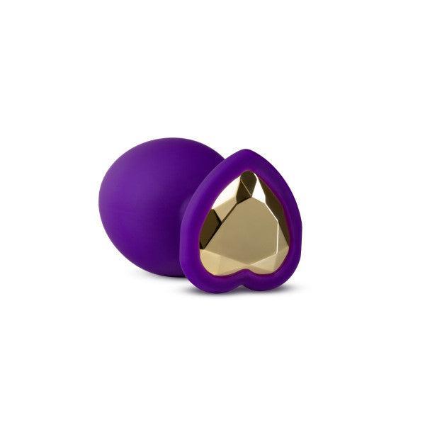 The Temptasia Bling Small Silicone Butt Plug in Purple by Blush features a small, heart-shaped design with a sparkling gold-colored heart embellishment on one end. Made from smooth silicone material, this butt plug is designed to stand upright on its heart-shaped base, making it ideal for beginner users.