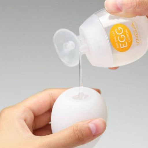 A person is holding a white, egg-shaped object with one hand and pouring gel from a small transparent bottle labeled "Tenga Egg Disposable Penis Masturbator - Lotion" with the other hand. This Tenga product offers a unique texture, ideal for those seeking something new. The background is plain and neutral.