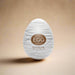 A white oval product with grey lines and a brown label reading "Tenga Egg Disposable Penis Masturbator - Silky 2" from Tenga, a New Adult Concept—your perfect travel buddy. This disposable masturbation sleeve is showcased against a plain white background.