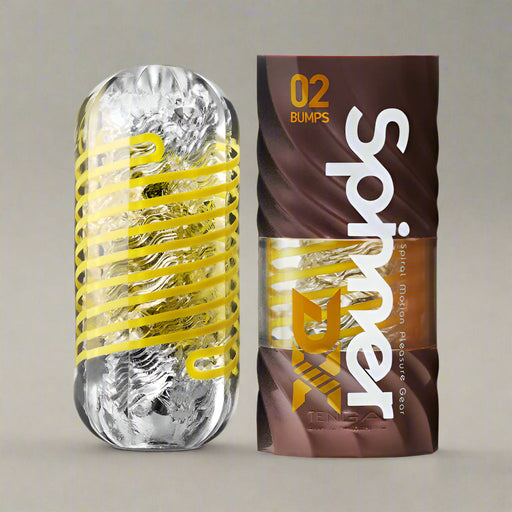 The Tenga Spinner DX Bumps Textured Masturbator, a transparent cylindrical device with a yellow spiral inside, stands beside its brown "02 Bumps" packaging featuring bold white "Spinner" text and vivid yellow geometric designs.