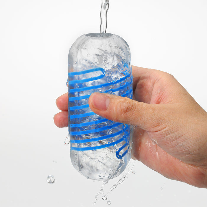 A hand holds a clear Tenga Spinner DX Drops Textured Masturbator with a blue spiral design as water is poured in, making the spinning stroker splash and overflow.