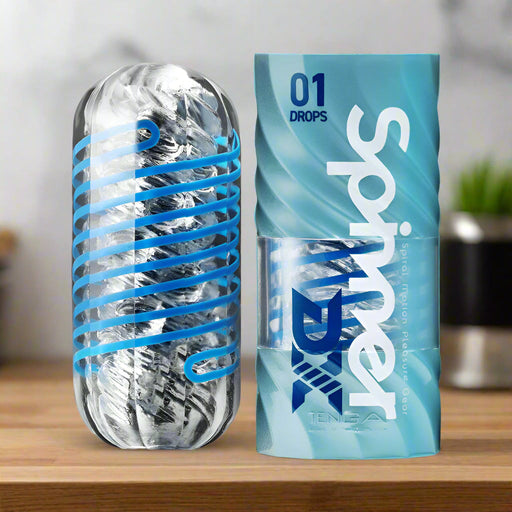 The Tenga Spinner DX Drops Textured Masturbator, featuring a clear capsule shape with silver and blue accents, stands by its blue "01 Drops Spinner" container, highlighting its thick elastomer for a unique spinning stroker experience.
