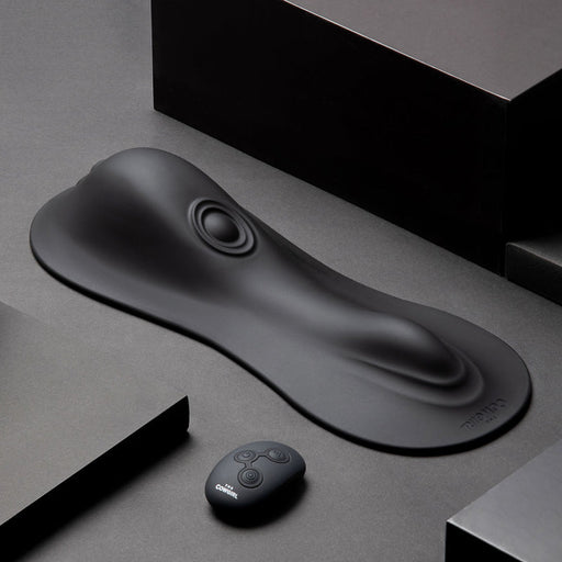 The Cowgirl Rodeo Rumbler Grinding Sex Machine features a sleek black silicone pad with raised contours and a circular indentation, plus a small remote control—ideal for hands-free stimulation—displayed on a dark geometric background.