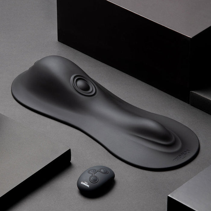 The Cowgirl Rodeo Rumbler Grinding Sex Machine features a sleek black silicone pad with raised contours and a circular indentation, plus a small remote control—ideal for hands-free stimulation—displayed on a dark geometric background.