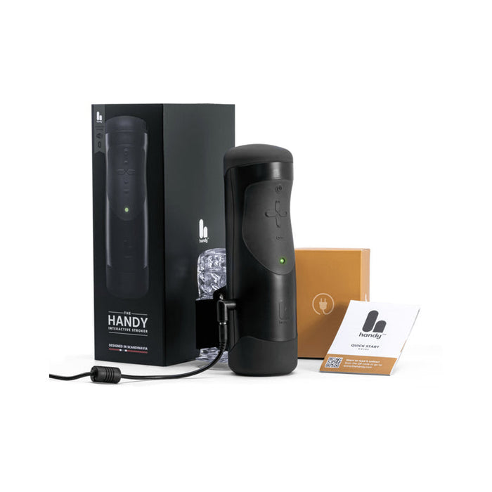 The Handy Adjustable Hand Job Penis Stroker in black, featuring a button and charging cord, is displayed on a white background with its packaging labeled "Handy," an instruction manual, and a small brown box.