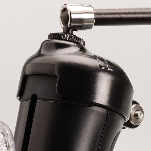 Close-up of The Handy Handsfree Cup Accessory, a black metallic device with a cylindrical body, handle attachment, shiny surface, visible screws and joints, and featuring a universal 1/4-inch screw against a light background.