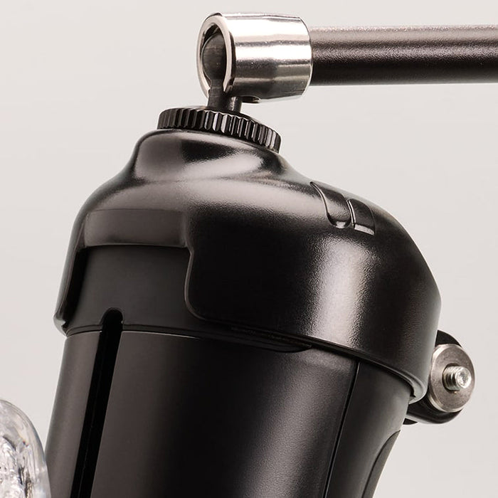 Close-up of The Handy Handsfree Cup Accessory, a black metallic device with a cylindrical body, handle attachment, shiny surface, visible screws and joints, and featuring a universal 1/4-inch screw against a light background.