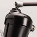 Close-up of The Handy Handsfree Cup Accessory, a black metallic device with a cylindrical body, handle attachment, shiny surface, visible screws and joints, and featuring a universal 1/4-inch screw against a light background.