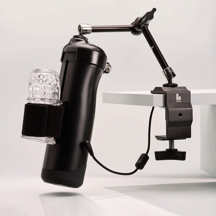 The Handy Handsfree Desk Mount features a black cylindrical device with a clear textured sleeve, secured by a strap and mounted to a white table with an articulating arm for adjustable positioning.