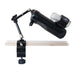 The Handy Handsfree Desk Mount, a black adjustable cup holder with an articulating arm, attaches to the edge of a light-colored wooden surface, securely holding a water bottle and a small clear container.