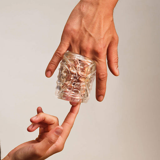 A clear, textured Handy Mermaid Collection Coral Sleeve - Intense Sensations, made from stretchy TPE, is wrapped around two fingers as another hand softly touches an exposed finger, all set against a neutral background.