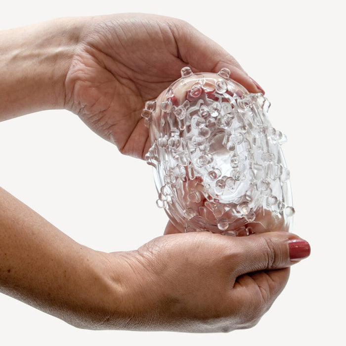 Two hands hold and gently squeeze The Handy TrueGrip Gen 2 Sleeve—a longer, thicker, stretchy, clear stroker sleeve with textured bumps—against a plain white background.