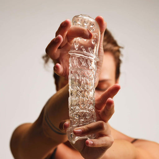 Someone stretches The Handy TrueGrip Gen 2 Sleeve—a longer, thicker, extra-stretchy transparent stroker with textured detailing and enhanced grip—while their face appears partially visible in the blurred background.