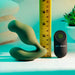 The Sergeant Powerful Prostate Vibrator with Remote Control - Green by Evolved Novelties, a curvy, green silicone remote control prostate vibrator, is displayed next to a ruler for scale. The black remote control features a distinctive green power button. The background includes folded towels and a small potted succulent to highlight the product's waterproof and submersible design alongside its 10 vibrating speeds.