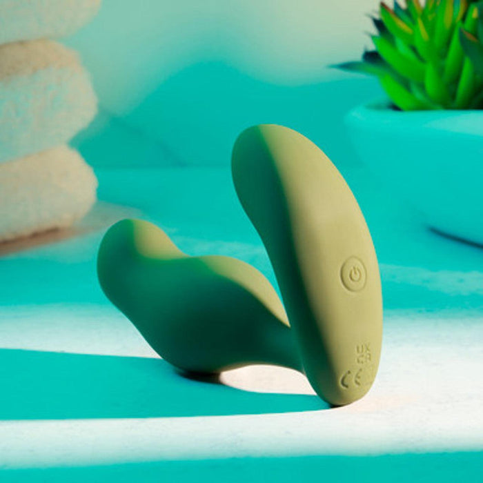 The Sergeant Powerful Prostate Vibrator with Remote Control by Evolved Novelties, a sleek green, dual-pronged device with a power button and certification marks, rests on a white surface under teal lighting. This waterproof and submersible vibrator offers 10 different vibrating speeds. In the background, there's a succulent plant in a white pot and folded towels stacked to the left.