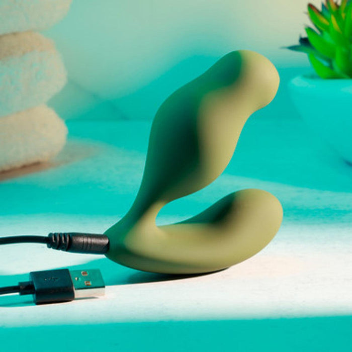 A green "Sergeant Powerful Prostate Vibrator with Remote Control" by Evolved Novelties, ergonomically designed and featuring dual ends with 10 vibrating speeds, lies charging via its USB cord on a light-colored surface. The background displays neatly stacked towels and a potted plant, all illuminated softly by teal light.