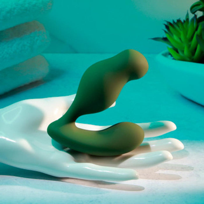 A green, abstract, S-shaped object, known as The Sergeant Powerful Prostate Vibrator with Remote Control - Green by Evolved Novelties, is displayed on a white human hand sculpture. The background features stacked towels and a potted succulent plant with a bluish-green hue, creating a serene and modern atmosphere. This waterproof and submersible item offers 10 vibrating speeds for the ultimate experience.
