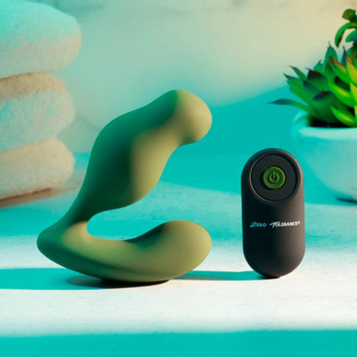 The Sergeant Powerful Prostate Vibrator with Remote Control - Green by Evolved Novelties, featuring 10 vibrating speeds and accompanied by a black remote control with a power button, rests on a white surface. In the background, slightly blurred, are stacked white towels and a potted plant. The scene is illuminated with teal lighting.
