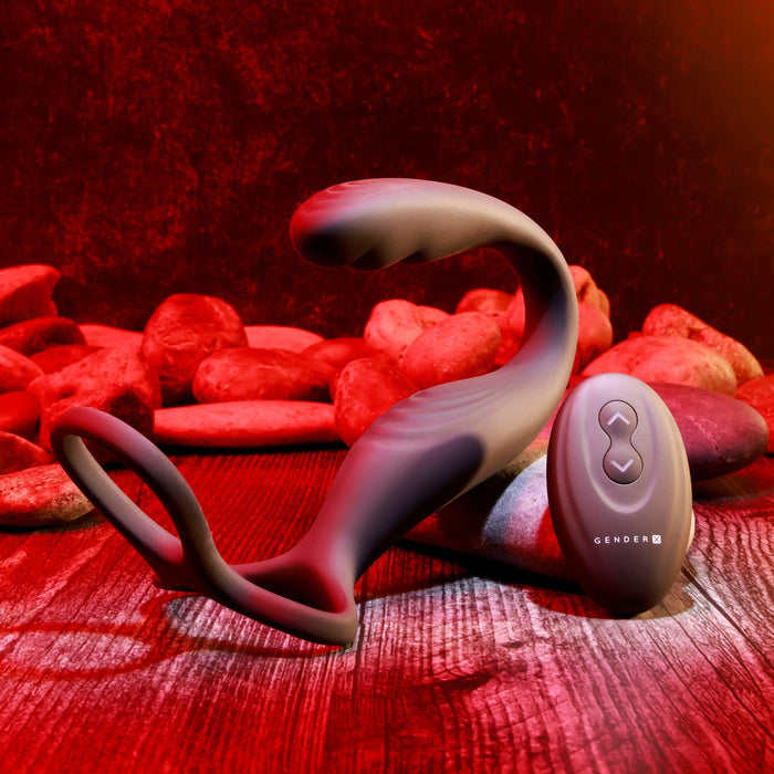 A black silicone sexual wellness device, identified as The Wrangler Vibrating Cock Ring and Butt Plug with Remote from Evolved Novelties, rests on a wooden surface surrounded by red stones. Boasting a smooth, ergonomic design with a ring at one end, it is accompanied by a remote control featuring two buttons and the brand name "Evolved Novelties." Bright red lighting accentuates the scene.