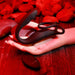 The Wrangler Vibrating Cock Ring and Butt Plug with Remote by Evolved Novelties, a black, dual-motor silicone product, is displayed on a prosthetic hand, resting on a surface surrounded by red stones. This waterproof adult toy is depicted under ambient red lighting.