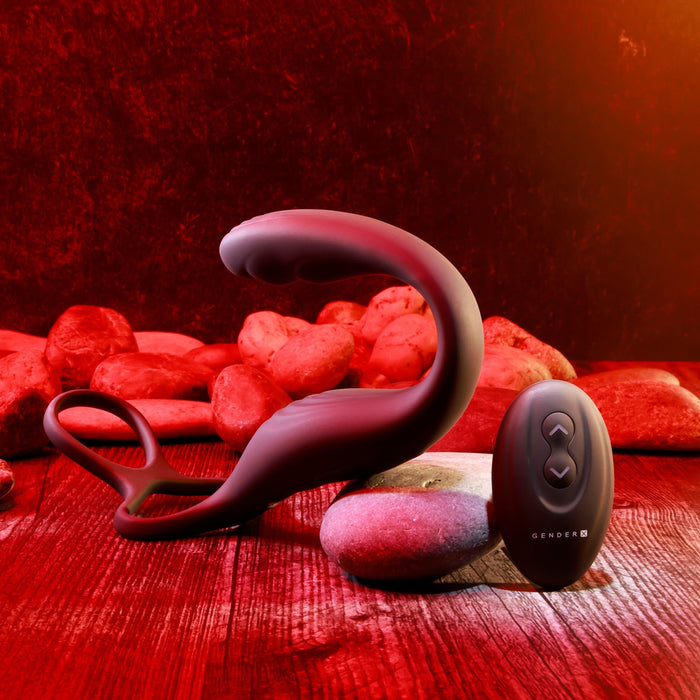 The Wrangler Vibrating Cock Ring and Butt Plug with Remote from Evolved Novelties, a sleek, black, curved waterproof sex toy with a looped handle, is elegantly displayed on wooden flooring adorned with red stones in the background. Accompanying it is a small matching remote control featuring three buttons. The scene is warmly lit with red and golden tones, highlighting the sophisticated design.