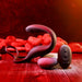 The Wrangler Vibrating Cock Ring and Butt Plug with Remote from Evolved Novelties, a sleek, black, curved waterproof sex toy with a looped handle, is elegantly displayed on wooden flooring adorned with red stones in the background. Accompanying it is a small matching remote control featuring three buttons. The scene is warmly lit with red and golden tones, highlighting the sophisticated design.