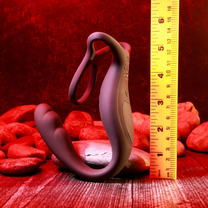 The Wrangler Vibrating Cock Ring and Butt Plug with Remote in black by Evolved Novelties is positioned vertically next to a yellow ruler, which shows that the device is approximately 6 inches tall. The background features red lighting and smooth stones on a wooden surface.