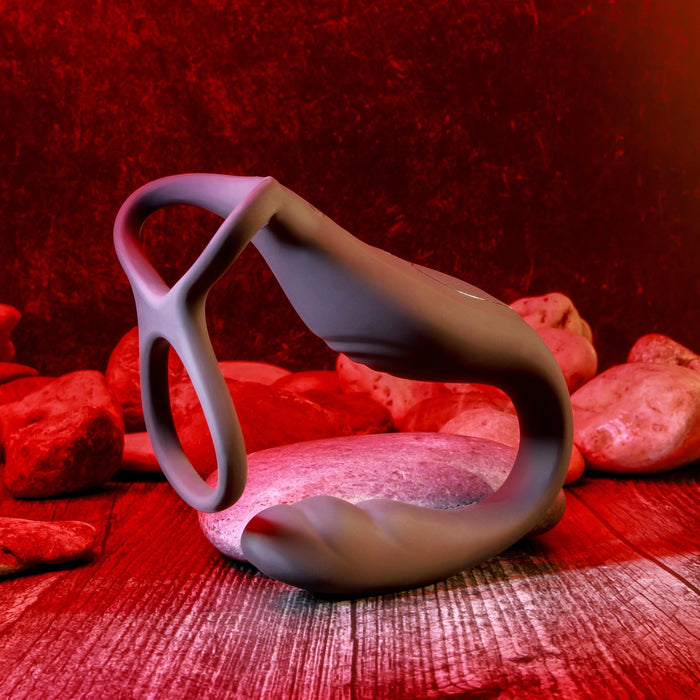 A sleek, contemporary sculpture with smooth, flowing lines is displayed on a circular stone, encircled by additional stones on a wooden base. The entire setting glows in red light, evoking an intense and dramatic feel akin to an Evolved Novelties advertisement for The Wrangler Vibrating Cock Ring and Butt Plug with Remote - Black.