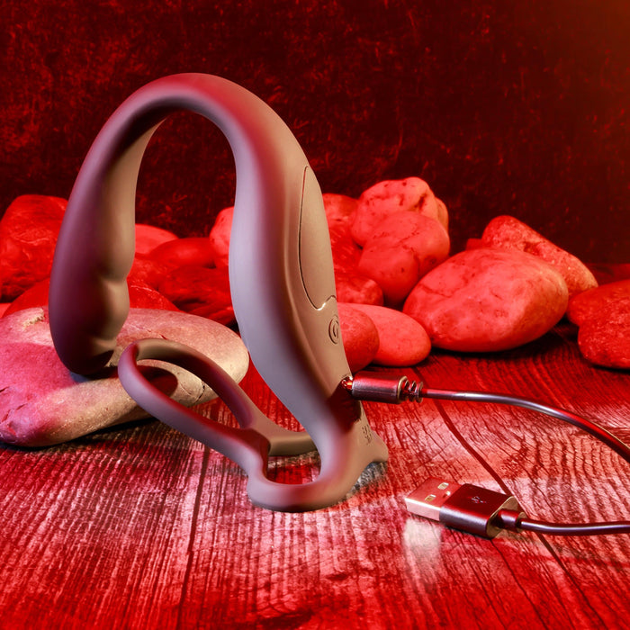 The Wrangler Vibrating Cock Ring and Butt Plug with Remote from Evolved Novelties is placed on a wooden surface with stones in the background. The setting, illuminated with red lighting, creates a warm atmosphere. The waterproof toy is connected to a USB charging cable.