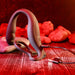 The Wrangler Vibrating Cock Ring and Butt Plug with Remote from Evolved Novelties is placed on a wooden surface with stones in the background. The setting, illuminated with red lighting, creates a warm atmosphere. The waterproof toy is connected to a USB charging cable.