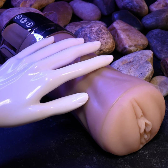 An XR Brands Tight Lipped Vibrating Vanilla Stroker with Suction + Free Movie Download with a flesh-toned exterior and a white robotic hand resting on it, set against a background of smooth round stones. The toy has a cylindrical shape with control buttons on its metallic top portion, offering varying vibrating speeds for enhanced pleasure.